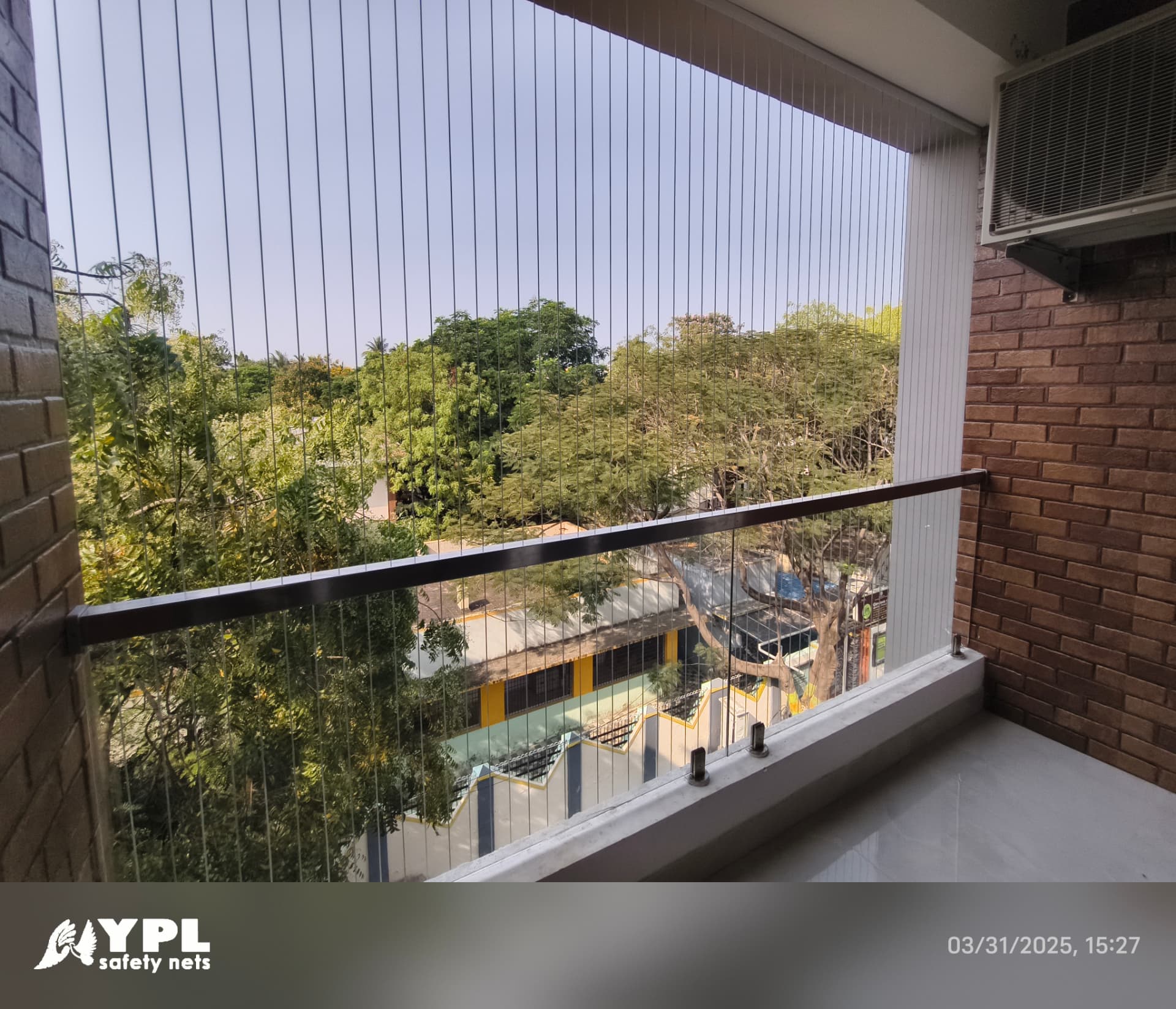 Balcony protected by vertical stainless steel invisible grill cables, with a glass railing and dark handrail, overlooking trees and neighbouring apartments in Chennai.