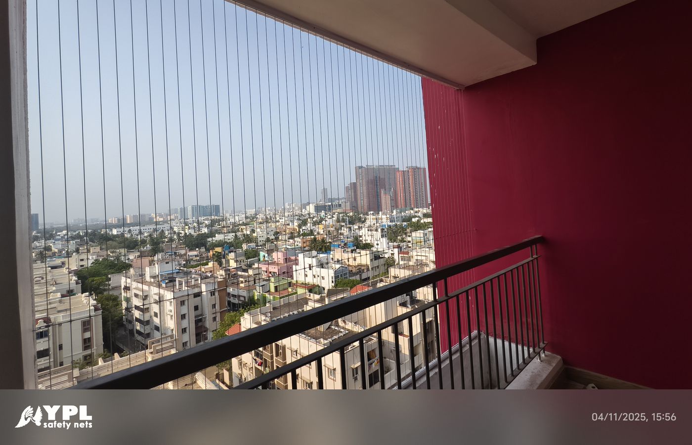 Vertical invisible grills enclose a high-rise balcony with a red wall and black railing overlooking dense city buildings.